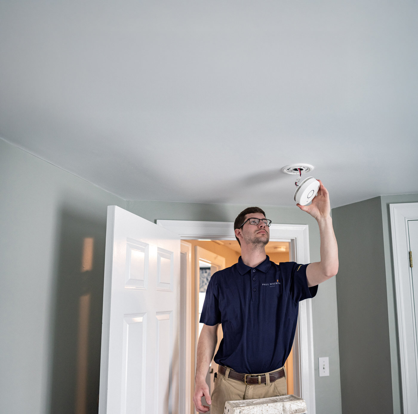 Paul Macrina electrician in black polo shirt installing white smoke detector on gray bedroom ceiling